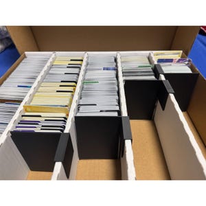 Pode incluir: Uma caixa de armazenamento de papelão marrom contendo várias fileiras de cartas colecionáveis organizadas com divisórias de plástico preto. As cartas estão empilhadas verticalmente em canais de papelão ondulado branco.
