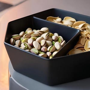 May include: A black, geometric serving bowl divided into three sections. One section is filled with shelled pistachios, another with pistachio shells, and the third is empty. The pistachios are green and tan.
