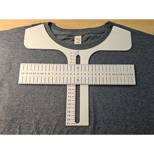 May include: A dark grey t-shirt featuring a white plastic t-shirt alignment tool placed over the chest area. The tool has a horizontal ruler marked from 0 to 38 cm on both sides and a vertical ruler marked from 0 to 53 cm.