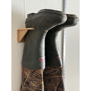 May include: A pair of dark green and camouflage patterned rubber boots hanging upside down on a beige wall-mounted plastic boot rack. The boots feature black textured soles and red text detailing near the top of the shaft, securely held in place.