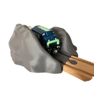 May include: A dark blue toy train engine with light green accents travelling through a grey plastic mountain tunnel piece. The tunnel is positioned over a section of wooden train track, creating a realistic railway scene for a toy train set.