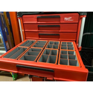 May include: A red Milwaukee Packout tool storage unit featuring three stacked drawers with black handles and a bottom drawer pulled open to reveal a series of grey plastic organisational bins with various compartment sizes for storage.