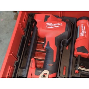 Puede incluir: Una clavadora inalámbrica Milwaukee roja y negra con un cargador lleno de clavos, guardada en un estuche de plástico rojo. La herramienta presenta el logotipo blanco de Milwaukee en el lateral y una empuñadura de goma negra para su manejo.
