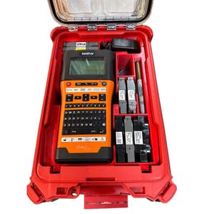Può includere: Un'etichettatrice Brother P-touch con scocca nera e arancione, contenuta in una valigetta rigida rossa. Il kit include diverse cartucce di nastro, un alimentatore e un pennarello, organizzati in scomparti sagomati su misura all'interno.