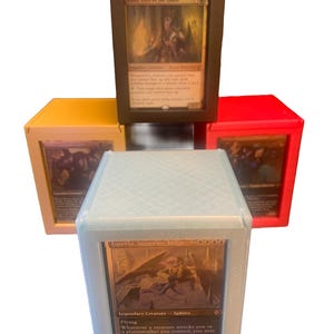 Op de afbeelding: Vijf kaartenopbergdozen in verschillende kleuren: lichtblauw, rood, geel en donkerbruin. Elke doos bevat een Magic: The Gathering-kaart. De kaarten tonen fantasy-kunst en tekst, waaronder "Isperia, Supreme Judge" en "Kaust, Eyes of the Glade".