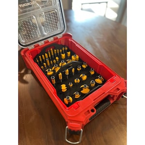 Puede incluir: Un maletín de almacenamiento de plástico rojo y negro con tapa transparente que muestra el texto PACKOUT, que contiene un inserto de espuma negra organizado con numerosas fresas de metal amarillo y plateado de diversas formas y tamaños.