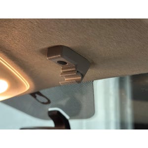 May include: A grey, L-shaped plastic clip mounted to the interior ceiling of a vehicle. The component features a flat base with a circular screw hole and a cylindrical, open-ended hook extending downwards, designed to secure a sun visor in place.