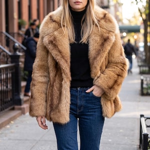 May include: A tan faux fur coat with long sleeves and a wide collar, worn over a black turtleneck jumper and dark blue denim jeans. The coat features a thick, textured pile and a straight hemline that reaches the upper hip area.