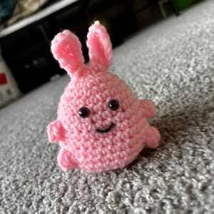 Op de afbeelding: Een kleine, roze gehaakte konijn met zwarte veiligheidsoogjes en een lachende mond. Het konijn heeft lange oren en kleine armen en benen. Het handgemaakte speelgoed ligt op een grijs tapijt.