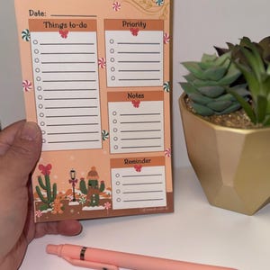 Könnte beinhalten: Ein pfirsichfarbener Notizblock mit Abschnitten für Things to-do, Priority, Notes und Reminder. Das Design zeigt Pfefferminzbonbons, rote Schleifen, einen Lebkuchenmann und einen Kaktus mit Wintermütze und Schal. Ein pfirsichfarbener Stift liegt darunter.