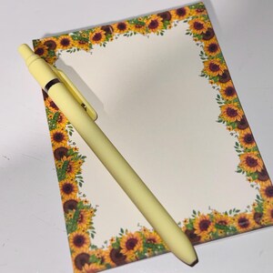 Könnte beinhalten: Ein gelber Stift liegt auf einem Notizblock mit einem Sonnenblumenrand. Der Notizblock hat leere Seiten, und das Sonnenblumenmuster weist gelbe, braune und grüne Farben auf. Der Stift ist ebenfalls gelb.