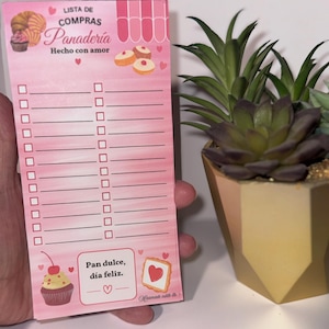 Może przedstawiać: Różowy notatnik z napisami "LISTA DE COMPRAS Panadería Hecho con amor" i "Pan dulce, día feliz." Notatnik zawiera ilustracje ciast i babeczek, z polami wyboru do listy zakupów.