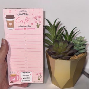 May include: A pink notepad with the words "LISTA DE COMPRAS Café y buena vibra" and a coffee cup illustration. A gold geometric planter holds several succulents. The notepad has a list format with checkboxes and a pink coffee cup illustration.
