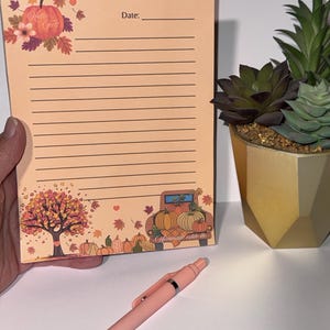 Puede incluir: Un bloc de notas con temática otoñal. El bloc de notas tiene un fondo melocotón con líneas horizontales para escribir. La parte superior del bloc de notas tiene las palabras "Hello Fall" y la palabra "Date:". La parte inferior del bloc de notas tiene un gráfico de un camión lleno de calabazas.