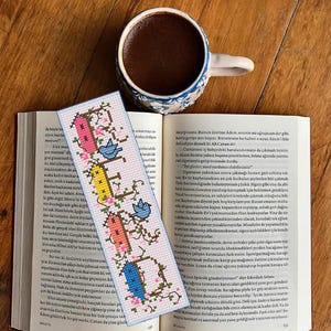 以下が含まれることがあります： ピンク、黄色、青の家をモチーフにした文字で「HOME」と縦に刺繍された長方形のクロスステッチのしおり。小さな青い鳥と緑の蔓が添えられており、コーヒーカップが置かれた開いた本の上に置かれています。