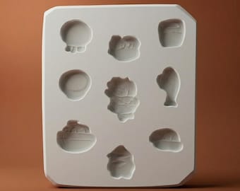 Puede incluir: Un molde de silicona blanco de forma rectangular que presenta ocho cavidades empotradas distintas con varias formas, incluyendo un barco, un pez y varias formas abstractas, diseñado para moldear materiales en figuras.
