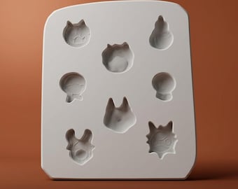 Puede incluir: Un molde de silicona blanco que presenta siete cavidades empotradas distintas con formas de varios rostros y figuras de personajes estilizados. El molde tiene una superficie lisa y plana con esquinas redondeadas para fundición.