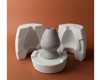 Puede incluir: Un jarrón de cerámica gris se encuentra sobre una base blanca, flanqueado por dos mitades de molde blancas. El jarrón tiene un cuerpo redondeado y un cuello estrecho. El fondo es marrón cálido liso.