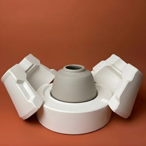 Puede incluir: Un jarrón de cerámica gris se encuentra en un molde blanco, con dos piezas de molde a cada lado. El jarrón tiene un cuerpo redondeado y un cuello estrecho. El fondo es de color naranja apagado.