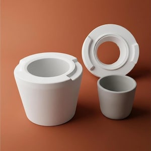 May include: Three ceramic pieces in shades of white and gray are arranged on a brown surface. The largest piece is a white, cylindrical container with a wide opening. A white circular piece and a small gray cup are also present.