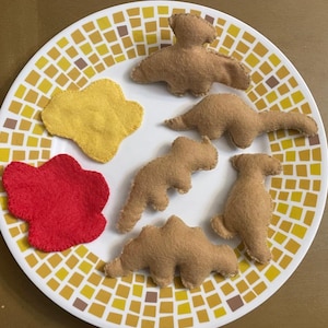 May include: A white plate with a yellow and brown mosaic pattern holds several felt dinosaur toys in shades of brown, yellow, and red. The toys are various shapes and sizes, including a stegosaurus and a pterodactyl.