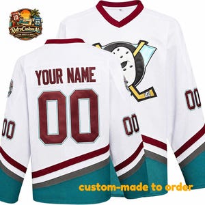 Puede incluir: Una camiseta de hockey blanca con cuello en V granate, rayas granates y verde azulado en las mangas y un gran logotipo de máscara de portero en el pecho. La parte posterior muestra el texto YOUR NAME sobre el número 00 en granate.