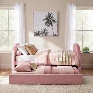 May include: A pink upholstered daybed with a pull-out trundle bed, featuring rounded headboard and footboard ends. The bed is dressed with white and pink bedding, including a textured throw blanket, several pillows, and a plush pink comforter.