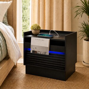 May include: Black nightstand with a built-in charging station and a magazine rack. A phone is charging on top. The magazine in the rack has the word "CERAE" on the cover. A small plant sits on top.