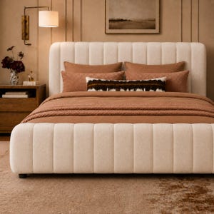 May include: A cream-colored upholstered bed frame with a vertical channel-tufted headboard and footboard. The bed is dressed with solid rust-colored bedding, two matching square pillows, and a long rectangular lumbar pillow with a dark brown pattern.