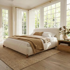 May include: A cozy bedroom scene featuring a cream-colored bed with a curved headboard, adorned with pillows and a brown blanket. A woven rug and wooden nightstand complete the inviting space. Large windows offer a view of the outdoors.