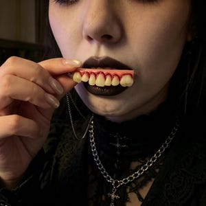 Può includere: Un accessorio protesico dentale caratterizzato da una fila di denti color bianco sporco fissati su una base rosa color gengiva con dettagli dipinti in rosso che imitano il sangue lungo il bordo gengivale, collegato a una sottile catena argentata.