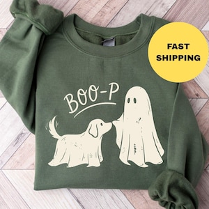 May include: An olive green long-sleeve sweatshirt featuring a cream-colored graphic of a dog and a ghost standing together. Above the figures, the text reads BOO-P in a simple font. The sweatshirt has a crew neckline and ribbed cuffs.