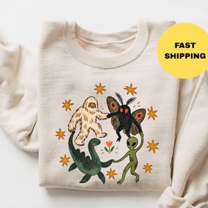 May include: A cream-colored crewneck sweatshirt featuring a graphic illustration of a beige Bigfoot, a black moth-like creature, a green sea monster, and a green alien holding hands in a circle around a small orange flower, surrounded by orange stars.