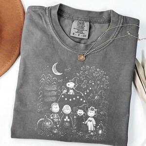 May include: A charcoal gray t-shirt featuring a white graphic print of Peanuts characters in Halloween costumes. The design includes Snoopy on a doghouse, pumpkins, a crescent moon, and bats. The shirt has a Comfort Colors label in size S.