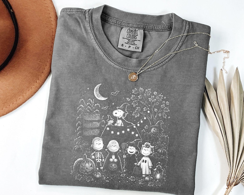 May include: A charcoal gray t-shirt featuring a white graphic print of Peanuts characters in Halloween costumes. The design includes Snoopy on a doghouse, pumpkins, a crescent moon, and bats. The shirt has a Comfort Colors label in size S.
