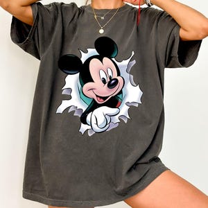 May include: A charcoal gray short-sleeve t-shirt featuring a graphic print of Mickey Mouse appearing to emerge through a torn white paper effect. The character is depicted with black ears, a peach-colored face, and white gloves, pointing forward.