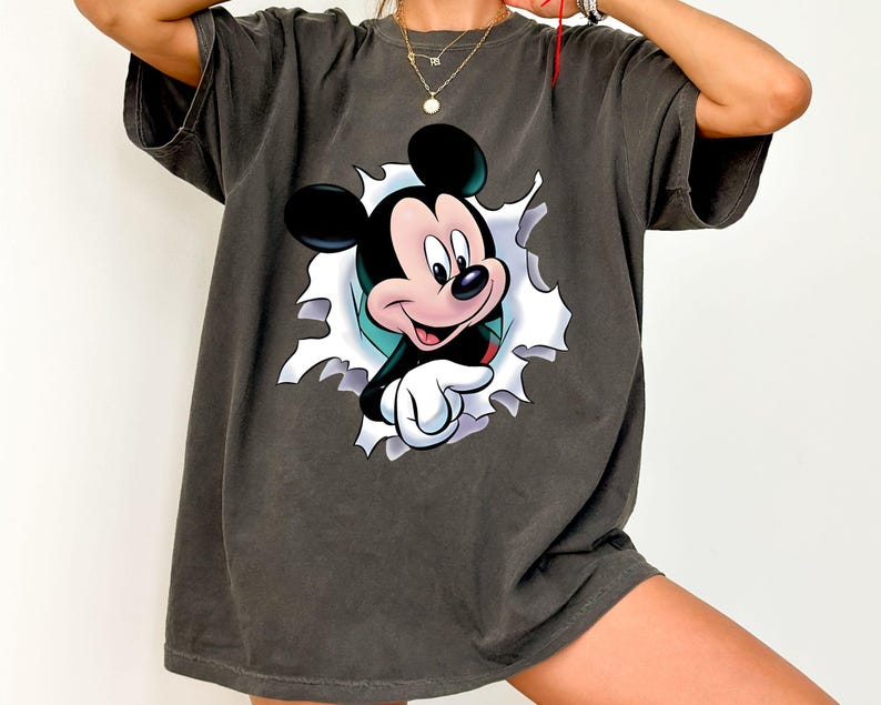 May include: A charcoal gray short-sleeve t-shirt featuring a graphic print of Mickey Mouse appearing to emerge through a torn white paper effect. The character is depicted with black ears, a peach-colored face, and white gloves, pointing forward.
