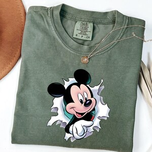 May include: An olive green short-sleeved t-shirt featuring a graphic of Mickey Mouse appearing to emerge from a torn white paper effect. The shirt has a crew neckline and a Comfort Colors brand label printed on the inside of the collar area.