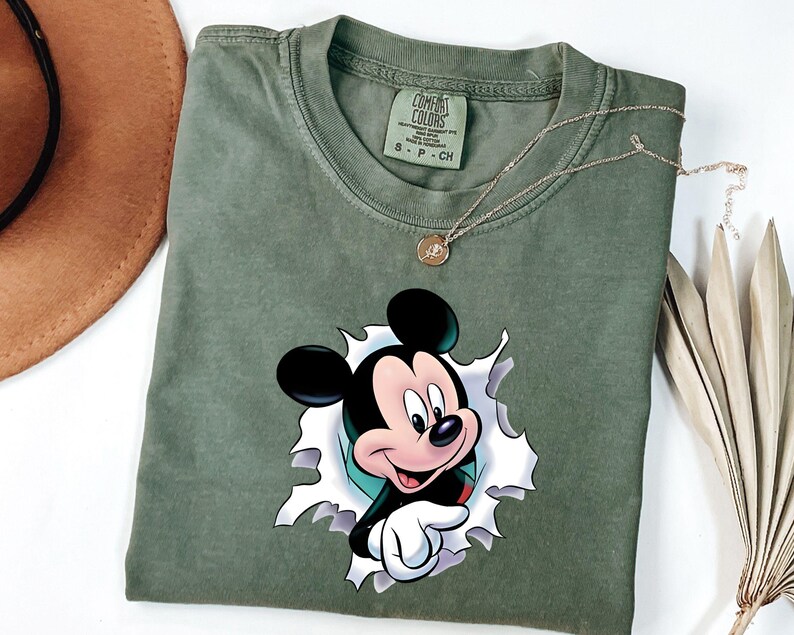 May include: An olive green short-sleeved t-shirt featuring a graphic of Mickey Mouse appearing to emerge from a torn white paper effect. The shirt has a crew neckline and a Comfort Colors brand label printed on the inside of the collar area.