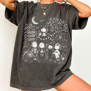 May include: A charcoal gray short-sleeve t-shirt featuring a white graphic print of Peanuts characters in Halloween costumes. The design includes a crescent moon, a tree, pumpkins, and signs reading PUMPKINS, HAY RIDES, and CORN MAZE.