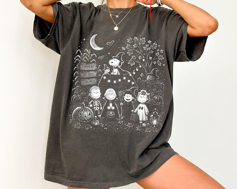 May include: A charcoal gray short-sleeve t-shirt featuring a white graphic print of Peanuts characters in Halloween costumes. The design includes a crescent moon, a tree, pumpkins, and signs reading PUMPKINS, HAY RIDES, and CORN MAZE.