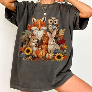 May include: A charcoal gray short-sleeve t-shirt featuring a central graphic of a fox, owl, squirrel, and rabbit surrounded by autumn leaves, sunflowers, and pumpkins. The animals and pumpkins have a textured, patchwork-style fabric pattern.