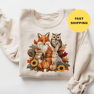 May include: A cream-colored crewneck sweatshirt featuring a central graphic of a fox, owl, squirrel, and rabbit surrounded by autumn leaves, sunflowers, and pumpkins. The design has a patchwork aesthetic. A yellow circle contains the text FAST SHIPPING.