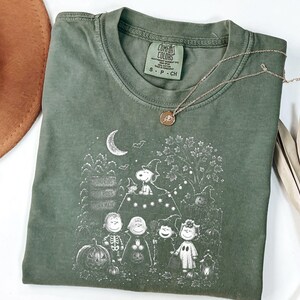 May include: An olive green t-shirt featuring a white graphic print of the Peanuts characters dressed in Halloween costumes. The design includes Snoopy, Charlie Brown, and friends near pumpkins, a crescent moon, and a sign. The shirt has a crew neck.