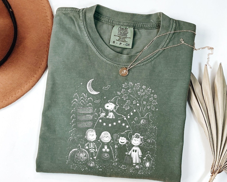 May include: An olive green t-shirt featuring a white graphic print of the Peanuts characters dressed in Halloween costumes. The design includes Snoopy, Charlie Brown, and friends near pumpkins, a crescent moon, and a sign. The shirt has a crew neck.