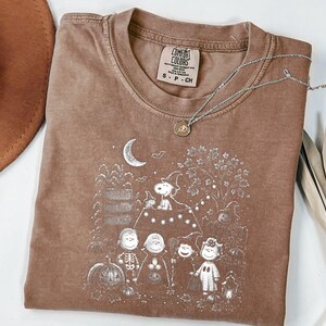 May include: A brown short-sleeve t-shirt featuring a white graphic print of Peanuts characters in Halloween costumes. The design includes Snoopy, Charlie Brown, and friends near a pumpkin patch, a crescent moon, and a signpost. The shirt has a crew neckline and a Comfort Colors brand label.
