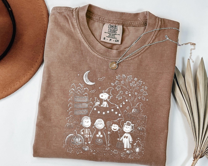 May include: A brown short-sleeve t-shirt featuring a white graphic print of Peanuts characters in Halloween costumes. The design includes Snoopy, Charlie Brown, and friends near a pumpkin patch, a crescent moon, and a signpost. The shirt has a crew neckline and a Comfort Colors brand label.