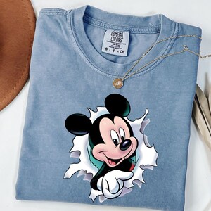 May include: A light blue short-sleeve t-shirt featuring a graphic of Mickey Mouse appearing to emerge through a torn opening. The shirt has a Comfort Colors brand label at the neck with size S printed on it. A thin gold chain necklace rests on top.