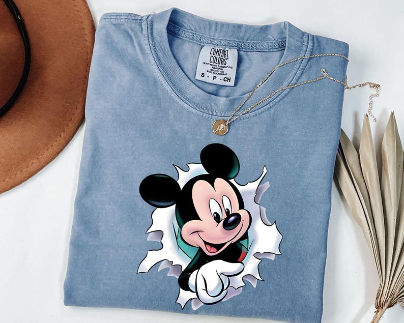 May include: A light blue short-sleeve t-shirt featuring a graphic of Mickey Mouse appearing to emerge through a torn opening. The shirt has a Comfort Colors brand label at the neck with size S printed on it. A thin gold chain necklace rests on top.