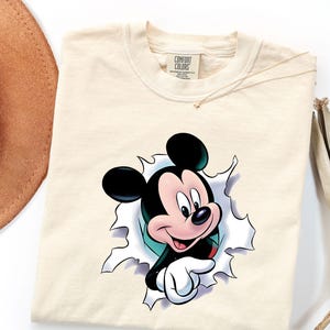 May include: A cream-colored t-shirt featuring a graphic of Mickey Mouse appearing to emerge from a torn paper effect. The character has black ears, a pink face, and white gloves. The shirt has a Comfort Colors brand label at the inner neckline.